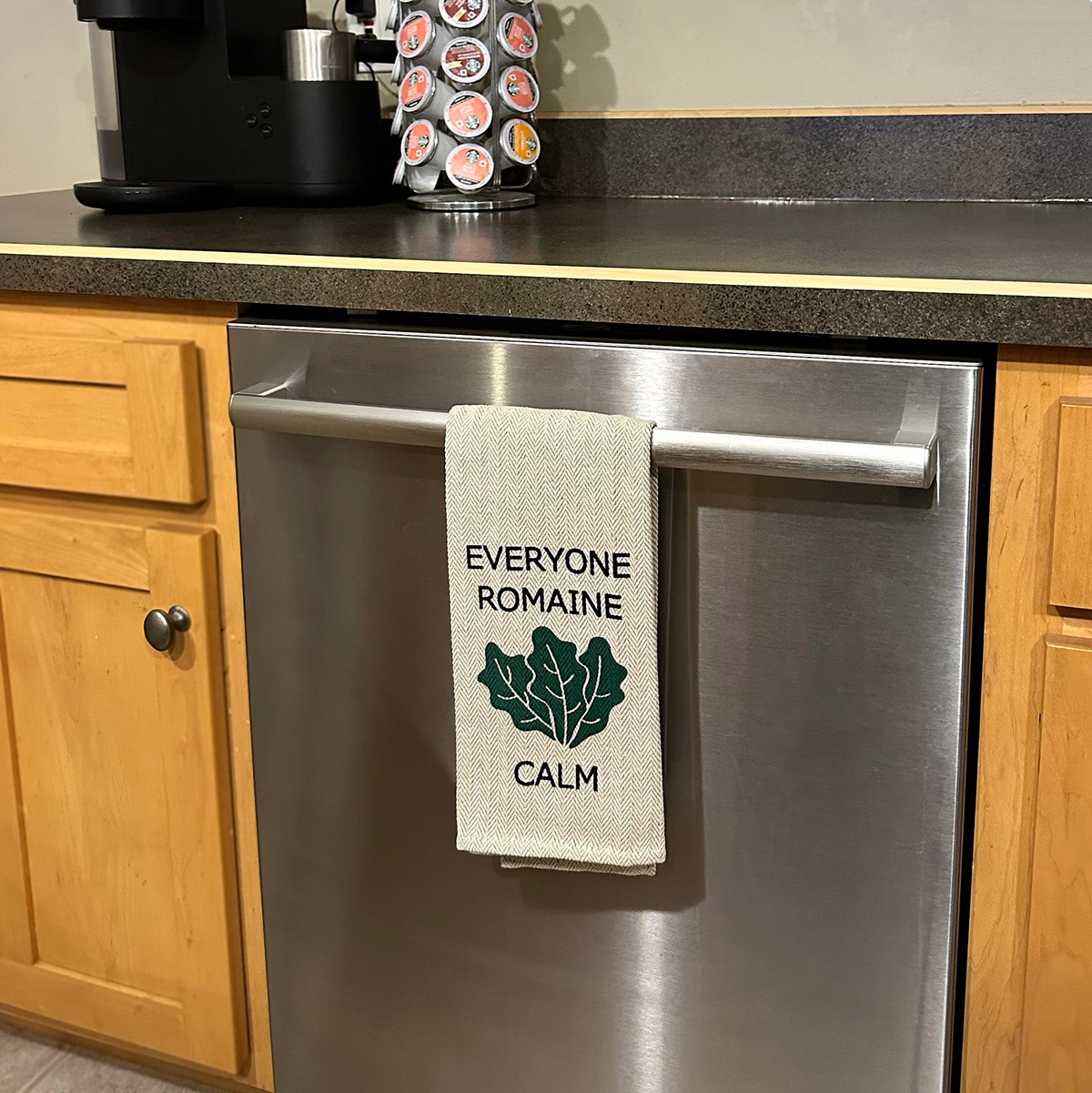 Kitchen Tea Towel with &quot;Everyone Romaine Calm&quot; design, neutral khaki color, hanging on a dishwasher handle.