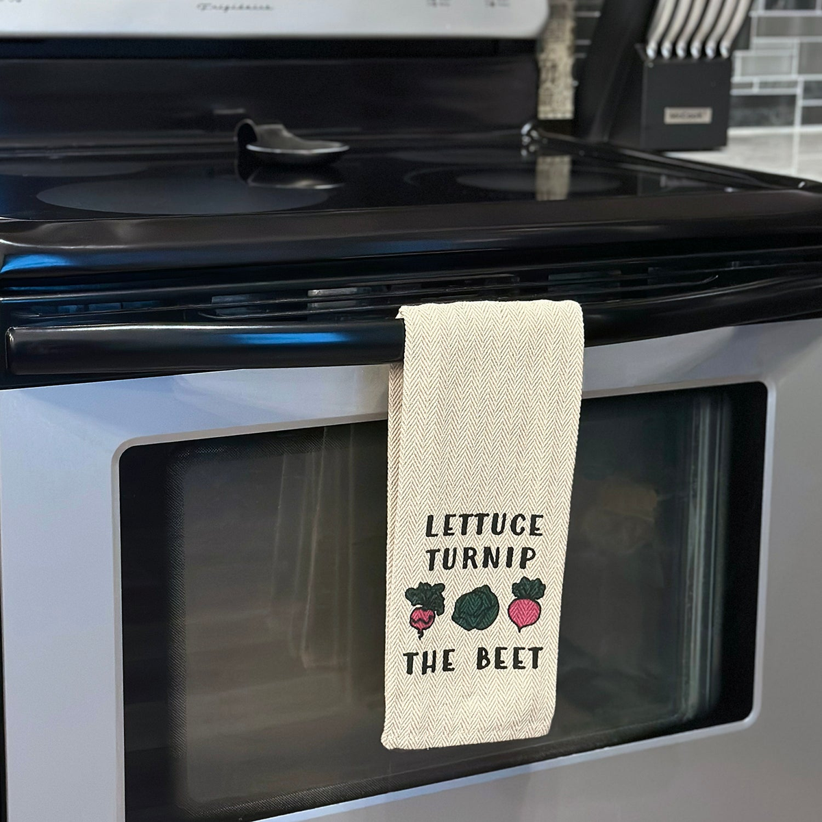 Kitchen tea towel with &quot;Lettuce Turnip the Beet&quot; design, 16x28 inches, hanging on oven.