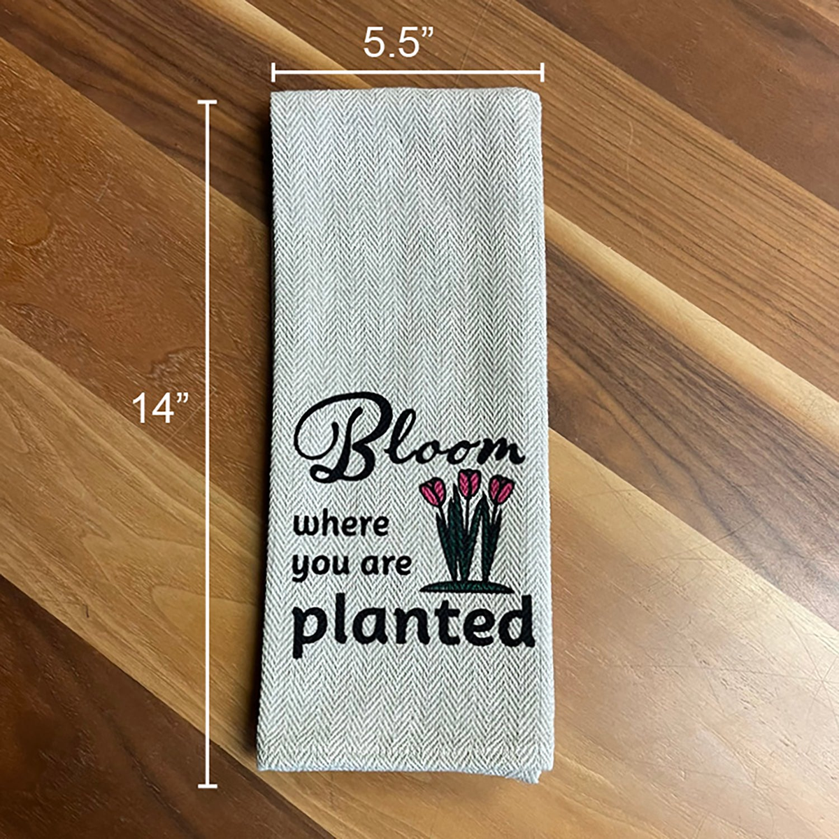 Kitchen Tea Towel folded on wooden surface, featuring &quot;Bloom Where You Are Planted&quot; design, size 5.5&quot; x 14&quot;.