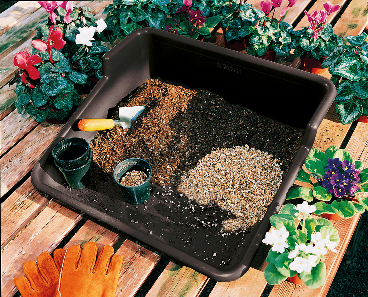 Black garden tidy tray with soil and seeds, featuring stainless steel trowel and cultivator with bamboo handles, surrounded by potted flowers.