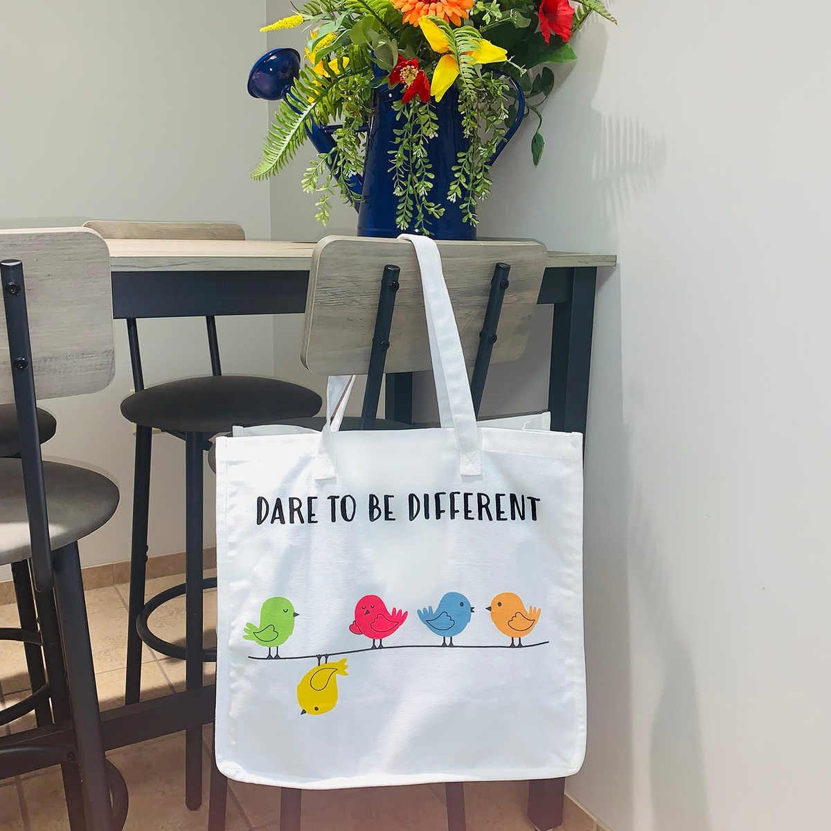 Bird themed cloth bag with colorful bird design and &quot;Dare to Be Different&quot; text, hanging on a chair.