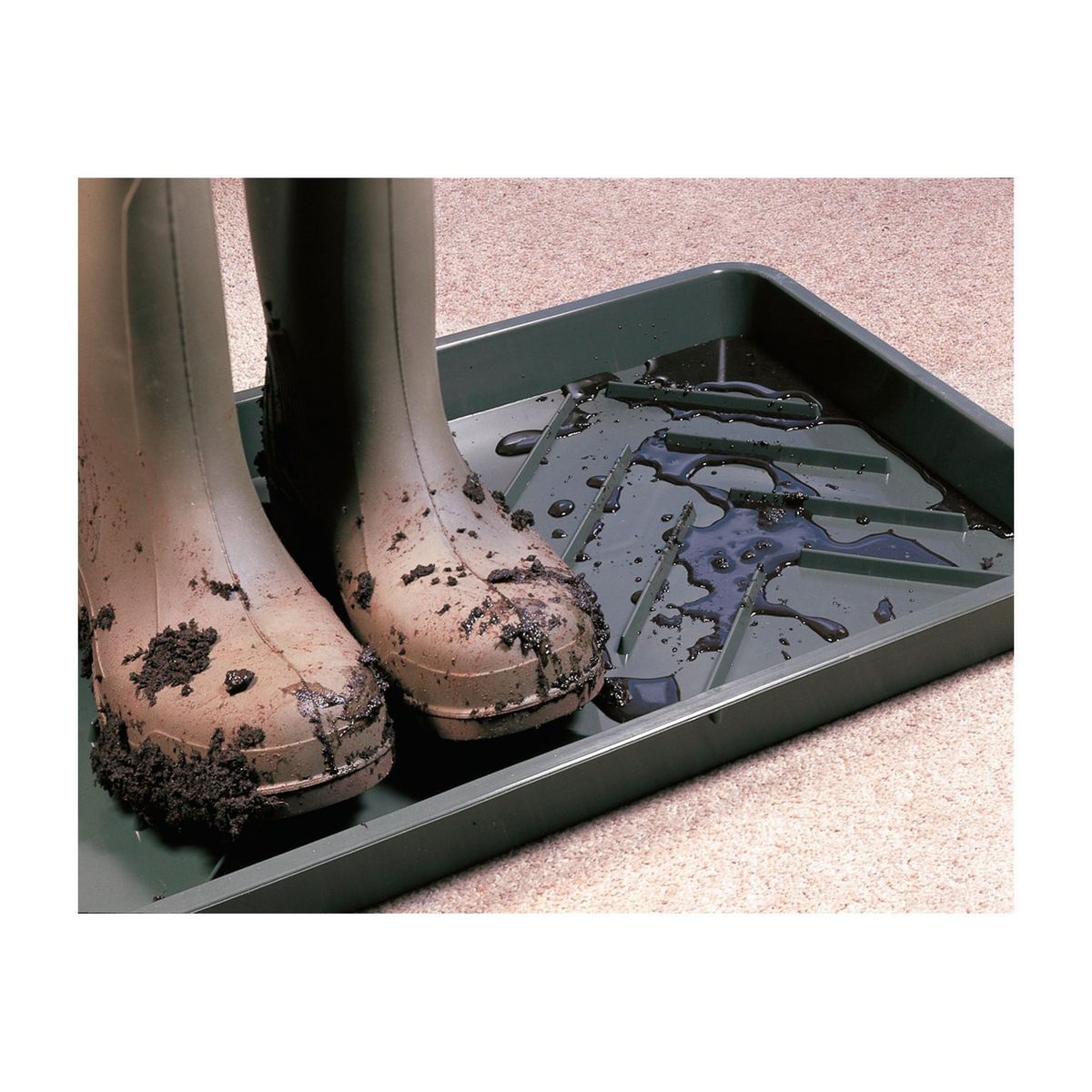 Black boot tray with raised ridges for mud scraping, shown with muddy boots on carpet.