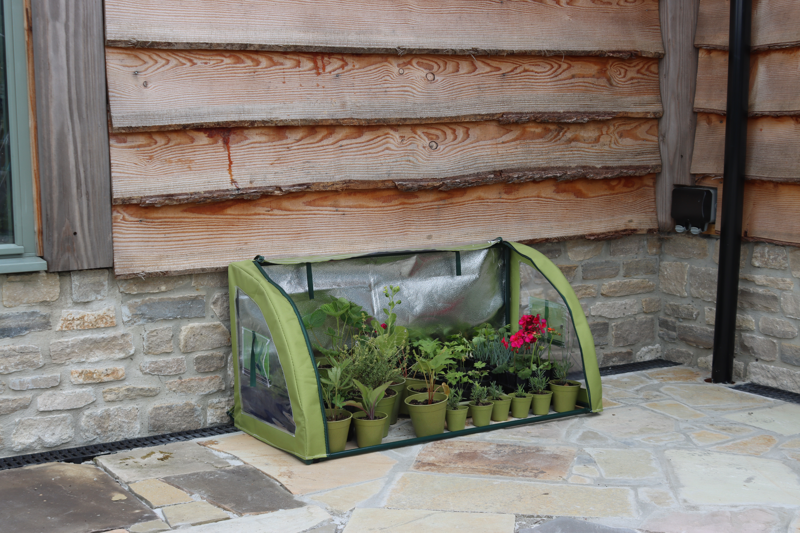 Light-Booster Coldframe with plants on a patio, featuring reflective backing and roll-up front panel.