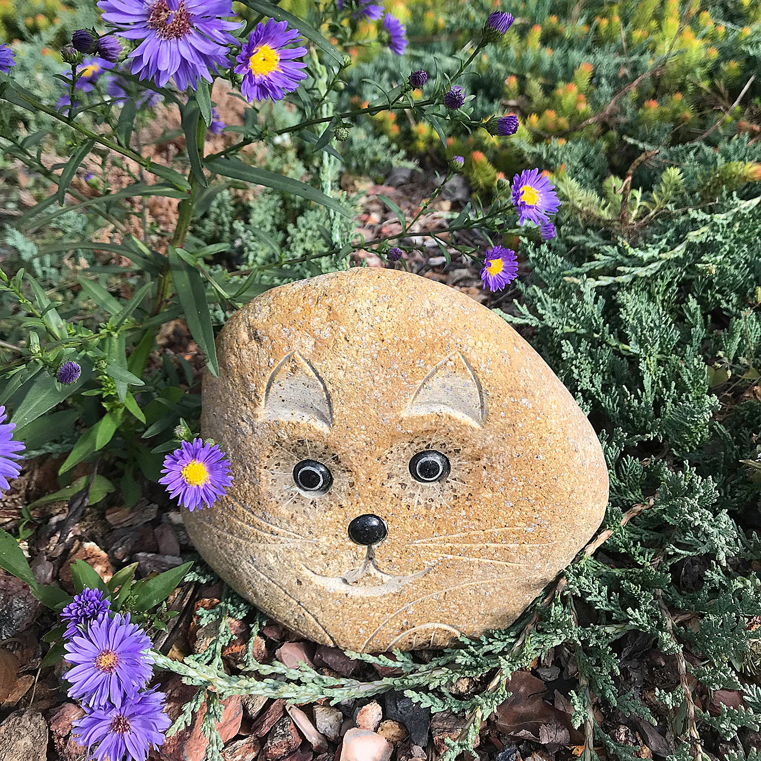 Hand carved natural granite boulder stone cat in garden setting with purple flowers.