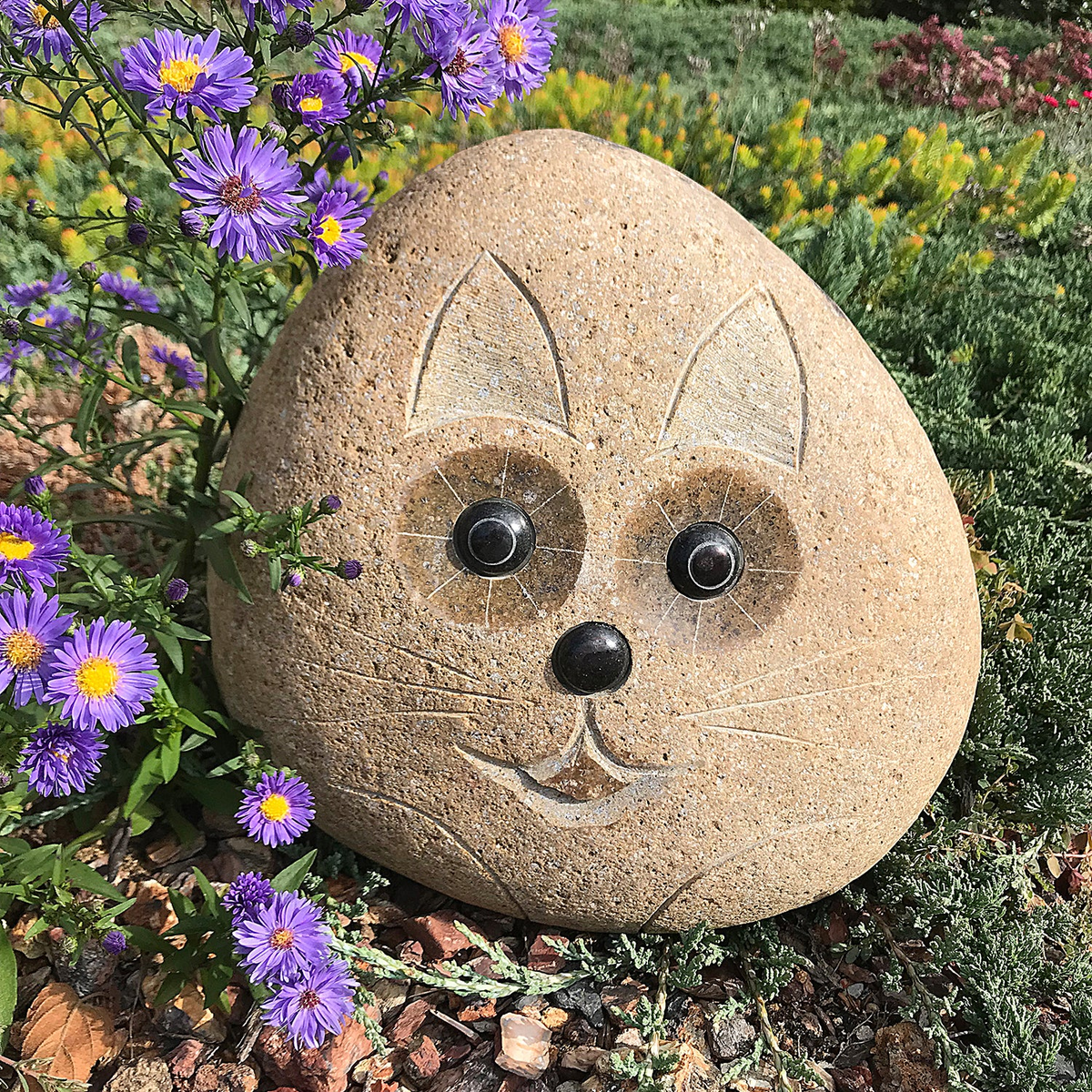 Hand carved natural granite boulder stone cat, 12&quot; tall, in a garden setting with flowers.