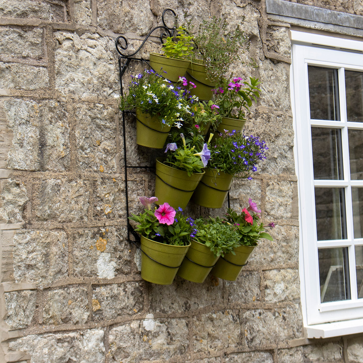 Wall plant holder with green pots against a stone wall displaying vibrant flowers and herbs.